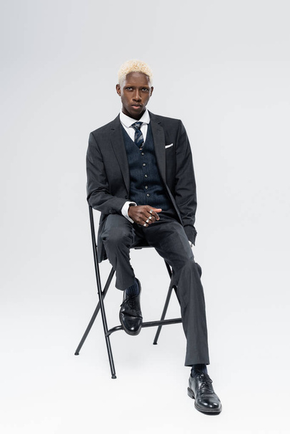 longitud completa de hombre afroamericano rubio en traje sentado en silla en gris  - Foto, Imagen