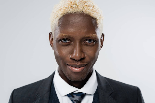 portrait of blonde and young african american man with blue eyes smiling isolated on grey  - Фото, изображение