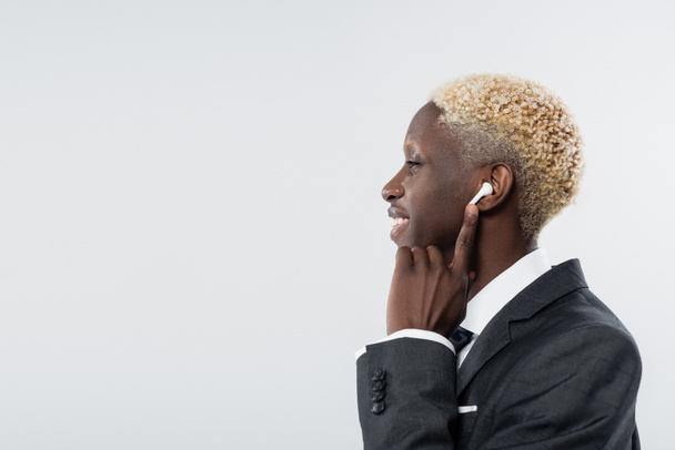 side view of happy african american man adjusting wireless earphone isolated on grey - Фото, зображення