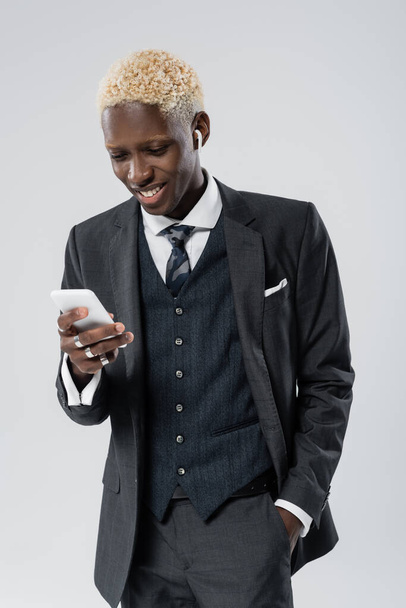 happy african american man in wireless earphone using smartphone isolated on grey - 写真・画像