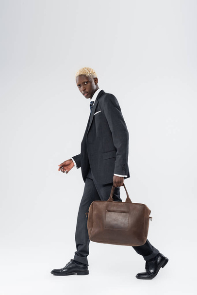 full length of blonde african american man in suit walking with leather bag on grey  - Foto, imagen