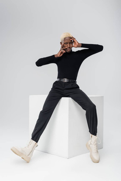 full length of young african american man in turtleneck touching face while posing on white cube on grey  - Foto, Imagen