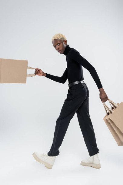 full length of stylish african american man in totally black outfit holding shopping bags on grey - Φωτογραφία, εικόνα