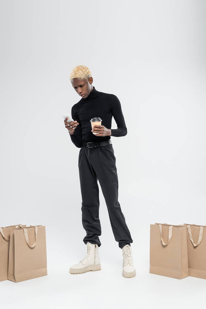piena lunghezza di uomo africano americano biondo in piedi con tazza di carta e smartphone vicino alle borse della spesa sul grigio  - Foto, immagini