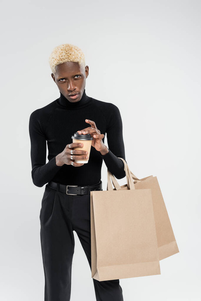 homem americano africano elegante de pé com sacos de compras e copo de papel isolado em cinza  - Foto, Imagem