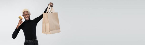 boldog afro-amerikai férfi gazdaság bevásárló táskák és hitelkártya elszigetelt szürke, banner - Fotó, kép