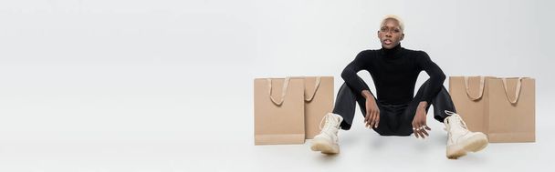 full length of blonde african american man sitting near paper bags on grey, banner - Фото, изображение