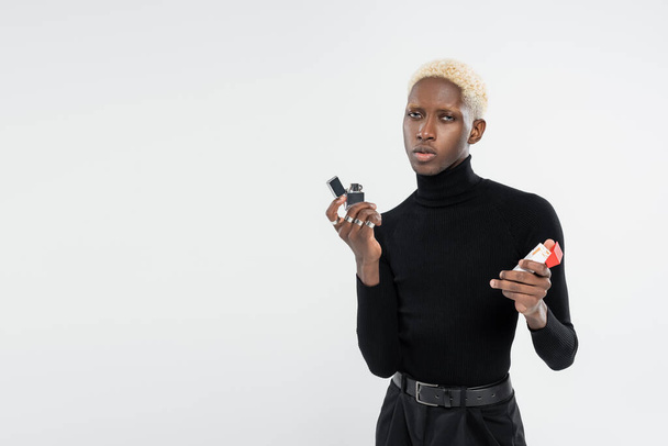 молодой и блондин африканский американец держит пачку сигарет и зажигалку изолированную на сером  - Фото, изображение