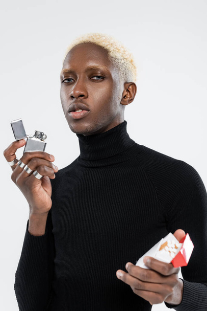 blonder afrikanisch-amerikanischer Mann hält Packung mit Zigaretten und Feuerzeug isoliert auf grau  - Foto, Bild