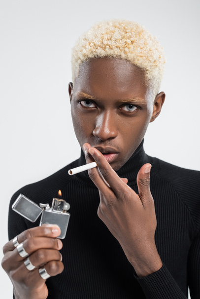 young and blonde african american man holding lighter with flame and cigarette isolated on grey  - Zdjęcie, obraz