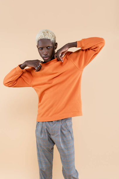 young african american man in orange sweatshirt looking at camera isolated on beige  - 写真・画像