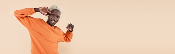 šťastný africký Američan v oranžové mikině při pohledu na kameru izolované na béžové, banner - Fotografie, Obrázek