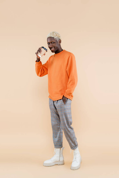 full length of blonde african american man in orange sweatshirt smiling while holding paper cup and posing on beige  - Fotoğraf, Görsel