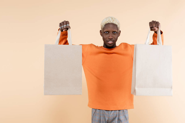 χαμογελαστός Αφροαμερικάνος με πορτοκαλί μπλούζα που κρατάει τσάντες για ψώνια απομονωμένες σε μπεζ - Φωτογραφία, εικόνα