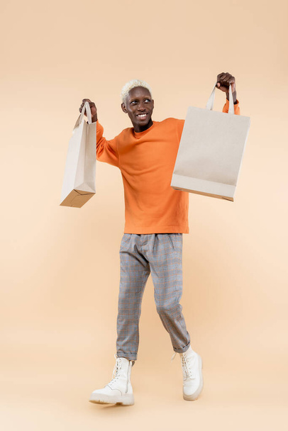 teljes hosszúságú boldog afro-amerikai férfi narancssárga pulóverben tartó bevásárló táskák bézs - Fotó, kép