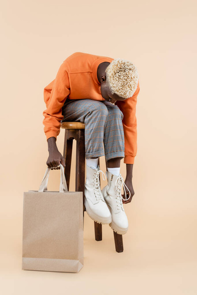 longitud completa de hombre afroamericano rubio en sudadera naranja con bolsa de compras y sentado en la silla en beige - Foto, Imagen
