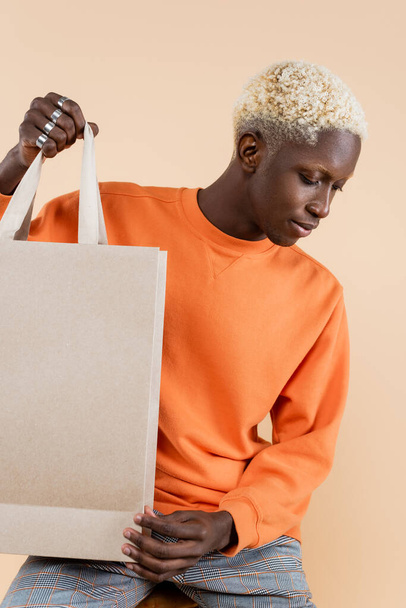 blonde african american man in sweatshirt holding shopping bag isolated on beige - Photo, Image
