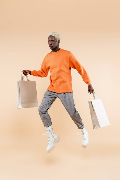 full length of blonde african american man in sweatshirt levitating while holding shopping bags on beige - 写真・画像