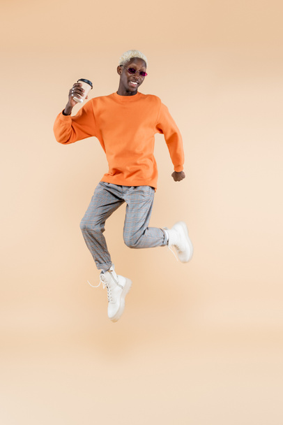 pleine longueur de heureux homme afro-américain en lunettes de soleil lévitant tout en tenant tasse en papier sur beige - Photo, image