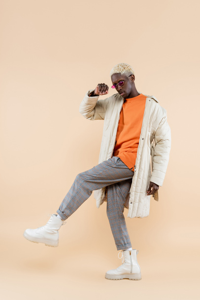 full length of young african american man in trendy sunglasses and coat posing on beige - Fotó, kép
