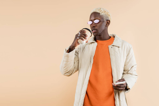 giovane uomo afroamericano in eleganti occhiali da sole e appendiabiti smartphone e coppa di carta isolati su beige - Foto, immagini