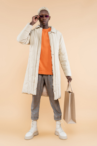 full length of young african american man in coat holding shopping bag while adjusting sunglasses on beige - Foto, afbeelding