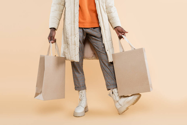 cropped view of african american man in coat holding shopping bags on beige - Fotoğraf, Görsel