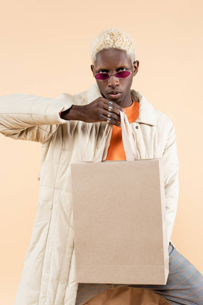 ξανθιά Αφροαμερικανός άνδρας με κομψό παλτό και μοντέρνα γυαλιά ηλίου κρατώντας τσάντα αγορών απομονωμένη σε μπεζ - Φωτογραφία, εικόνα