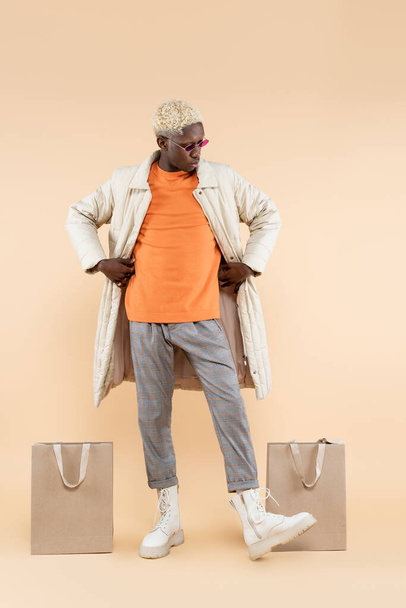 piena lunghezza dell'uomo afroamericano alla moda in elegante cappotto e occhiali da sole in posa vicino a borse della spesa sul beige - Foto, immagini
