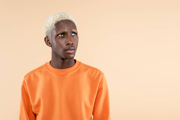 dreamy african american man in orange sweatshirt looking away isolated on beige  - Photo, Image