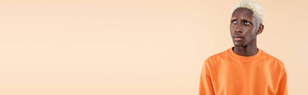 zasněný africký Američan v oranžové mikině odvrací pohled izolovaný na béžové, banner  - Fotografie, Obrázek