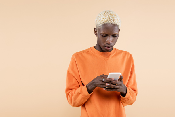 blonde african american man in orange sweatshirt using smartphone isolated on beige  - Fotó, kép