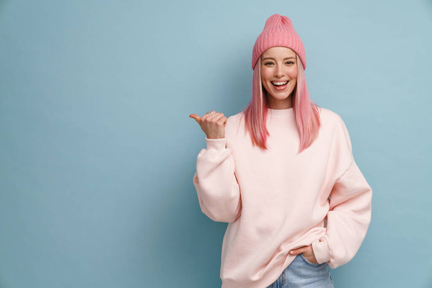 Young woman wearing pink hat smiling and pointing finger aside isolated over blue wall - Photo, Image