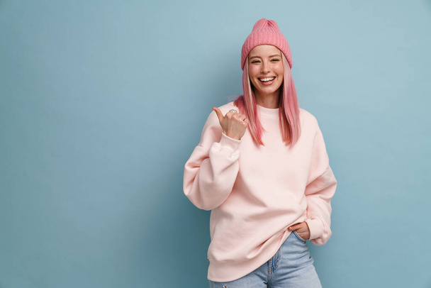 Young woman wearing pink hat laughing and pointing finger aside isolated over blue wall - Fotografie, Obrázek