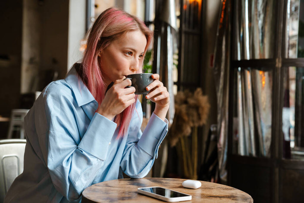 Young white woman with pink hair drinking coffee while sitting in cafe - Foto, afbeelding