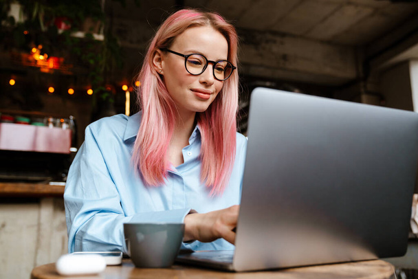 Young smiling woman wearing eyeglasses working with laptop while sitting in cafe - Foto, immagini