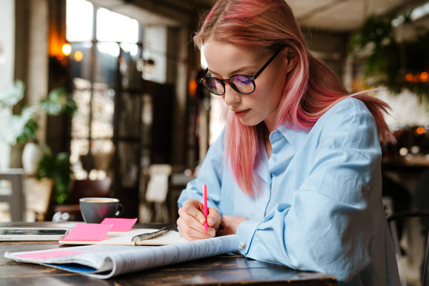 Fiatal gyönyörű nő rózsaszín hajjal tanul és jegyzetel, miközben ül a kávézóban - Fotó, kép
