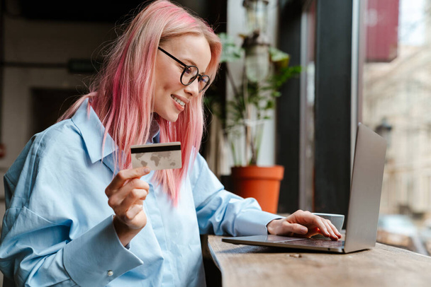 Young beautiful woman with pink hair using laptop and credit card while sitting in cafe - Foto, Imagem