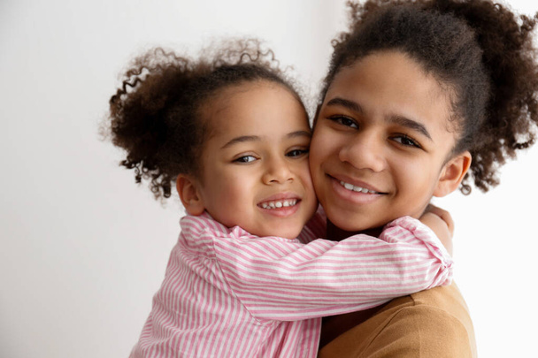 Younger and older sister spending time together at home. Two black girls of different age hugging and showing affection. Black female siblings having fun and bonding. Background, copy space, close up. - 写真・画像