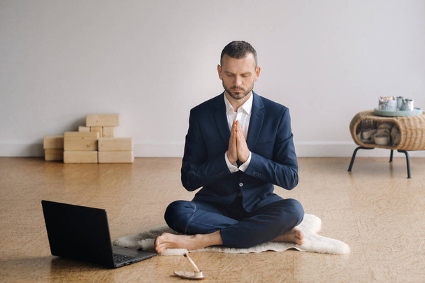 A man in a formal suit meditates while sitting in a fitness room with a laptop - Фото, зображення