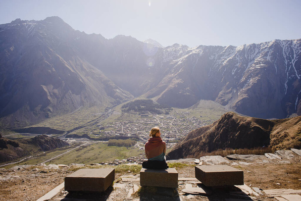 A girl with long hair is sitting with her back against the backdrop of a mountain landscape with snow-capped peaks and a village below - Photo, Image