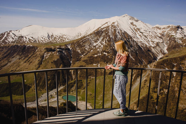 A girl with long hair stands against the backdrop of a mountain landscape with snow-capped peaks and a beautiful lake below - Fotoğraf, Görsel