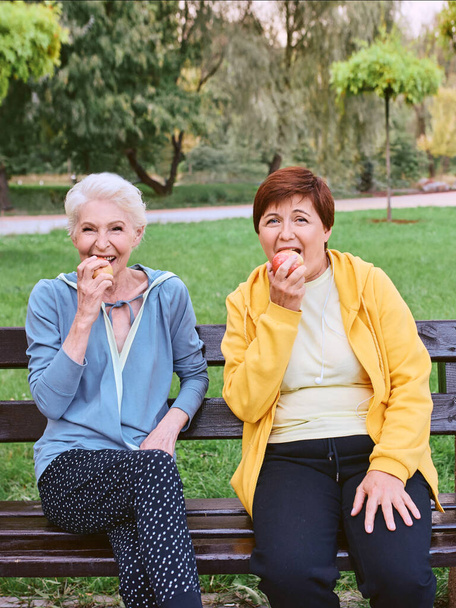 dwie dojrzałe kobiety jedzące jabłka na ławce po ćwiczeniach sportowych w parku. koncepcja zdrowego stylu życia - Zdjęcie, obraz