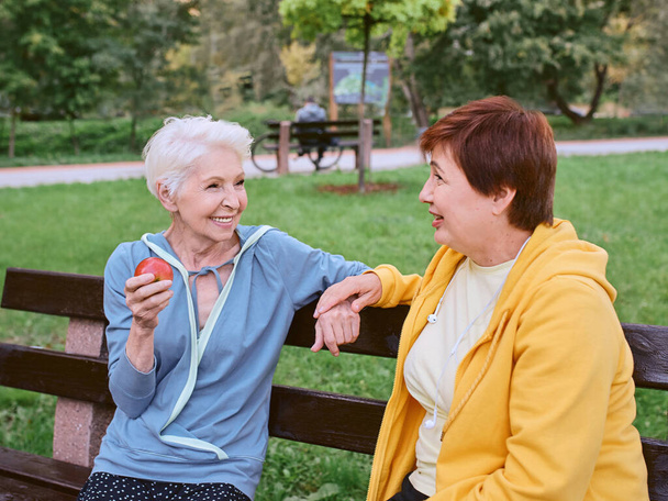 Két érett nő almát eszik a padon, miután a parkban tornáztak. egészséges életmód koncepció - Fotó, kép