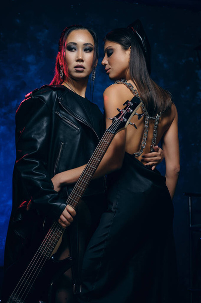 Two beautiful young women wear fashion clothes posing with guitar indoors, studio shot. Fashion and beauty concept - Foto, Imagen