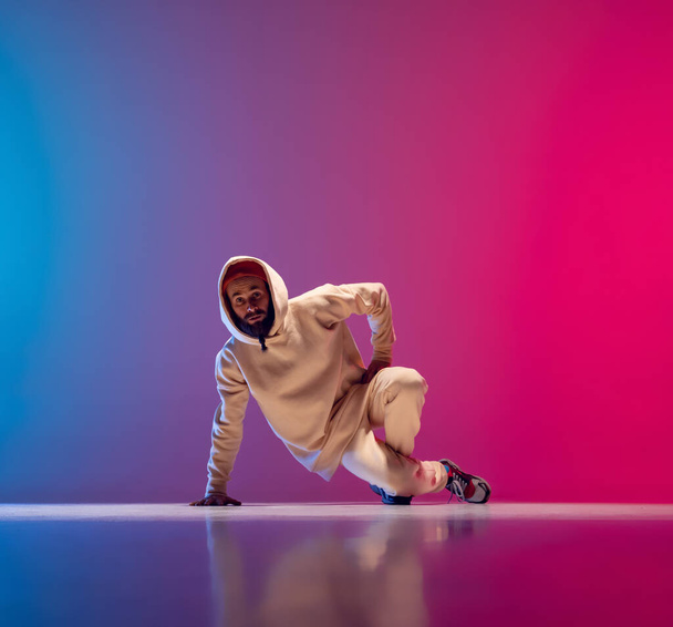 Studio shot of young flexible sportive man dancing breakdance in white outfit on gradient pink blue background. Concept of action, art, beauty, sport, youth - Fotó, kép