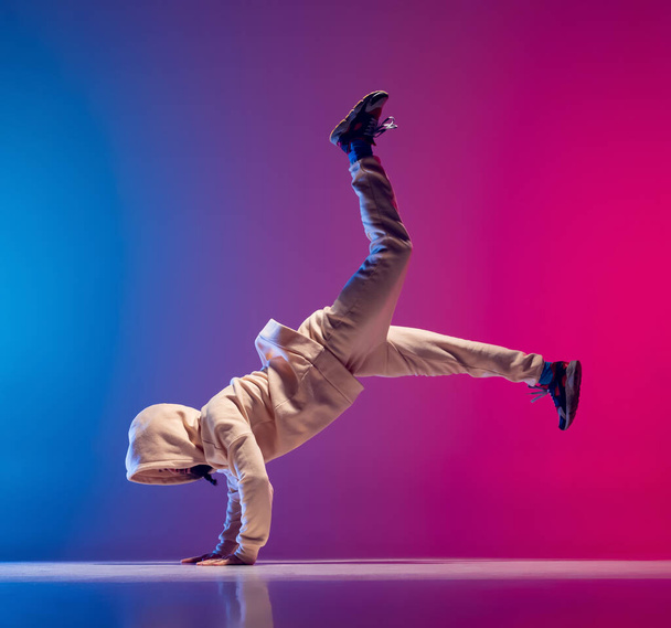 Studio shot of young flexible sportive man dancing breakdance in white outfit on gradient pink blue background. Concept of action, art, beauty, sport, youth - Фото, изображение