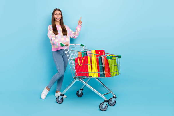 Photo of confident lady hold trolley direct finger empty space wear sweater jeans sneakers isolated blue color background - Foto, Imagem