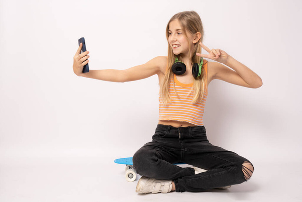 Πλήρες μήκος πορτρέτο όμορφο νεαρό κορίτσι σε casual ρούχα κάθεται στο skateboard χρησιμοποιώντας smartphone απομονωμένο σε λευκό φόντο. Έννοια τρόπου ζωής ανθρώπων - Φωτογραφία, εικόνα