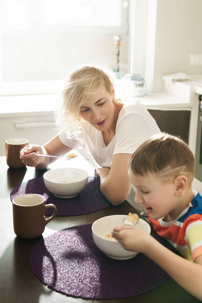 Beautiful mother and her cute son eating healthy cornflakes for breakfast at the sunny morning - 写真・画像